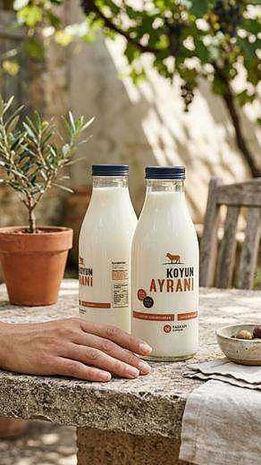 Two Taşkapı Çiftliği ayran bottles on a stone courtyard table with an olive plant and a bowl of olives